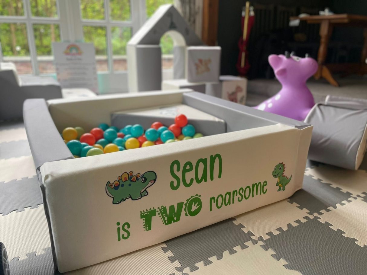 Two 'Roarsome' dinosaur-themed soft play birthday setup with custom vinyl, dinosaur hoppers, ride-on toys, and scattered decals.