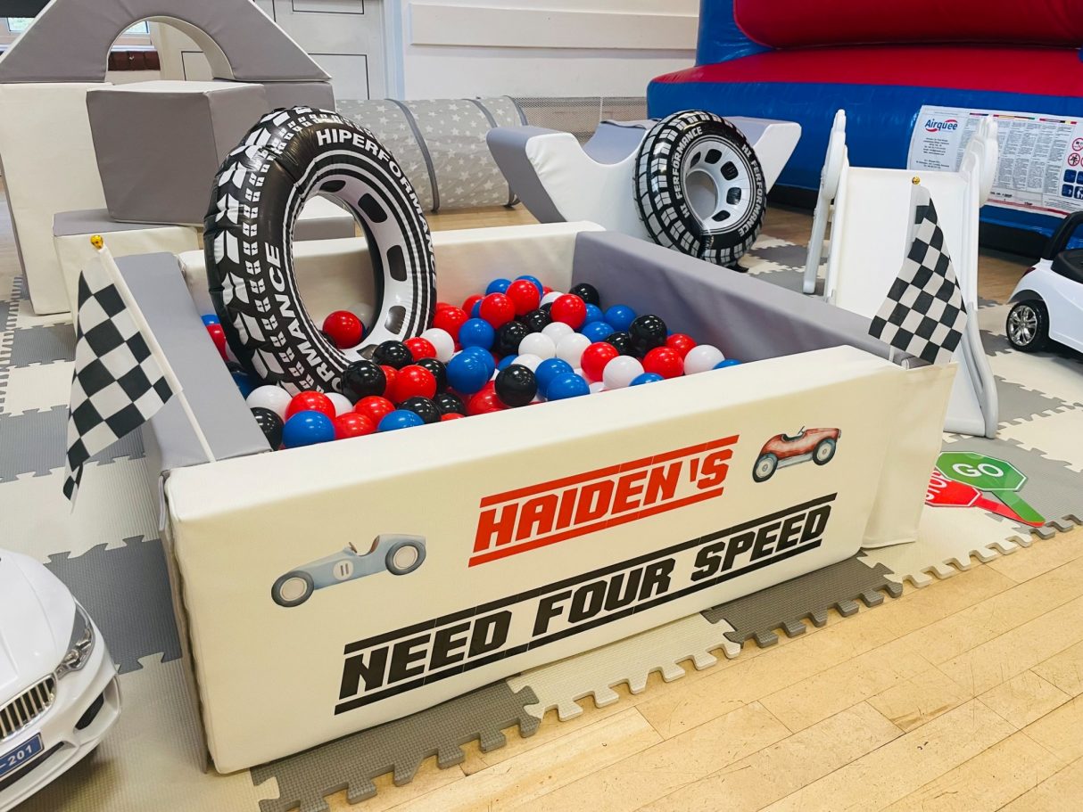Racing themed setup featuring a personalised ball pit with checkered flags, inflatable tyres, and red, black, and white balls. 