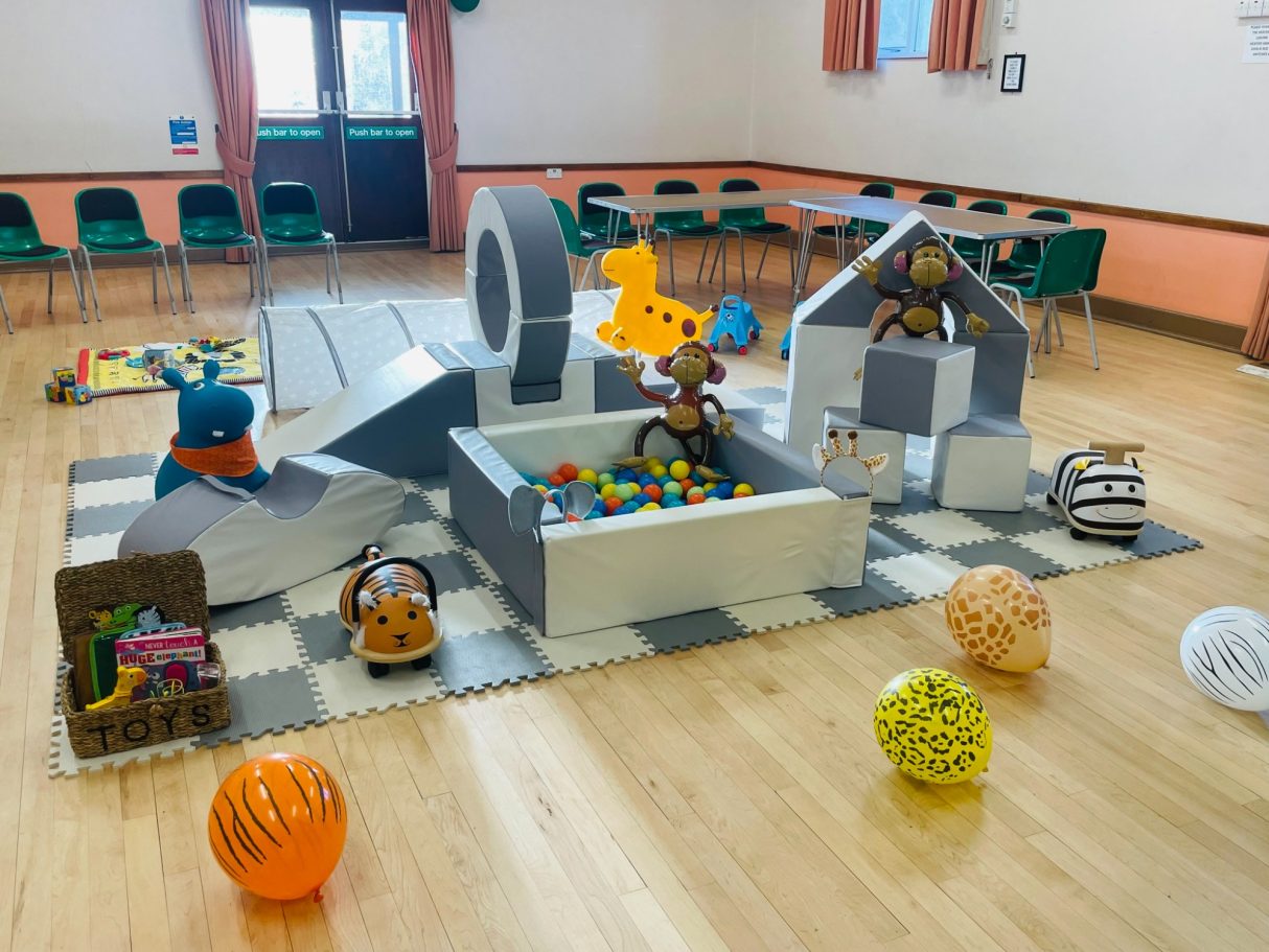 Colourful play area with soft toys, play structures, and a ball pit on a floor mat.